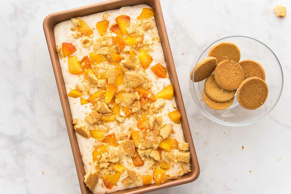 A loaf pan of easy peach cobbler ice cream being assembled. A bowl of vanilla wafers is set to the right.
