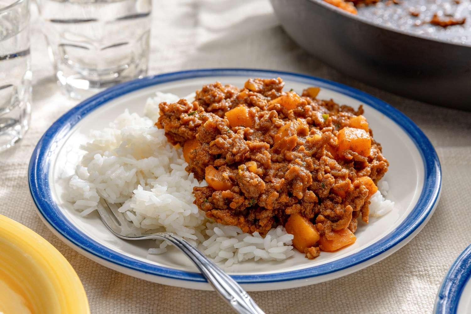 Angled view of a white plate with a blue rim of picadillo over rice on a white table cloth