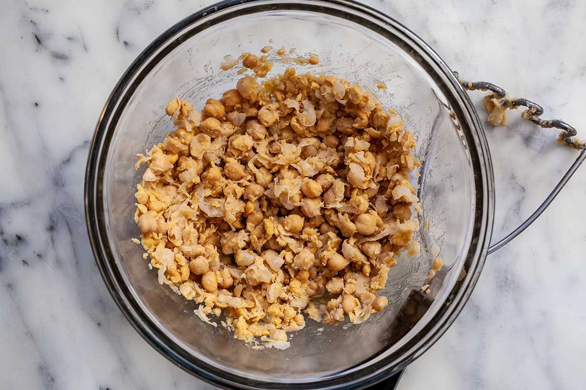 Canned chickpeas are mashed in a glass bowl with a potato masher next to it.