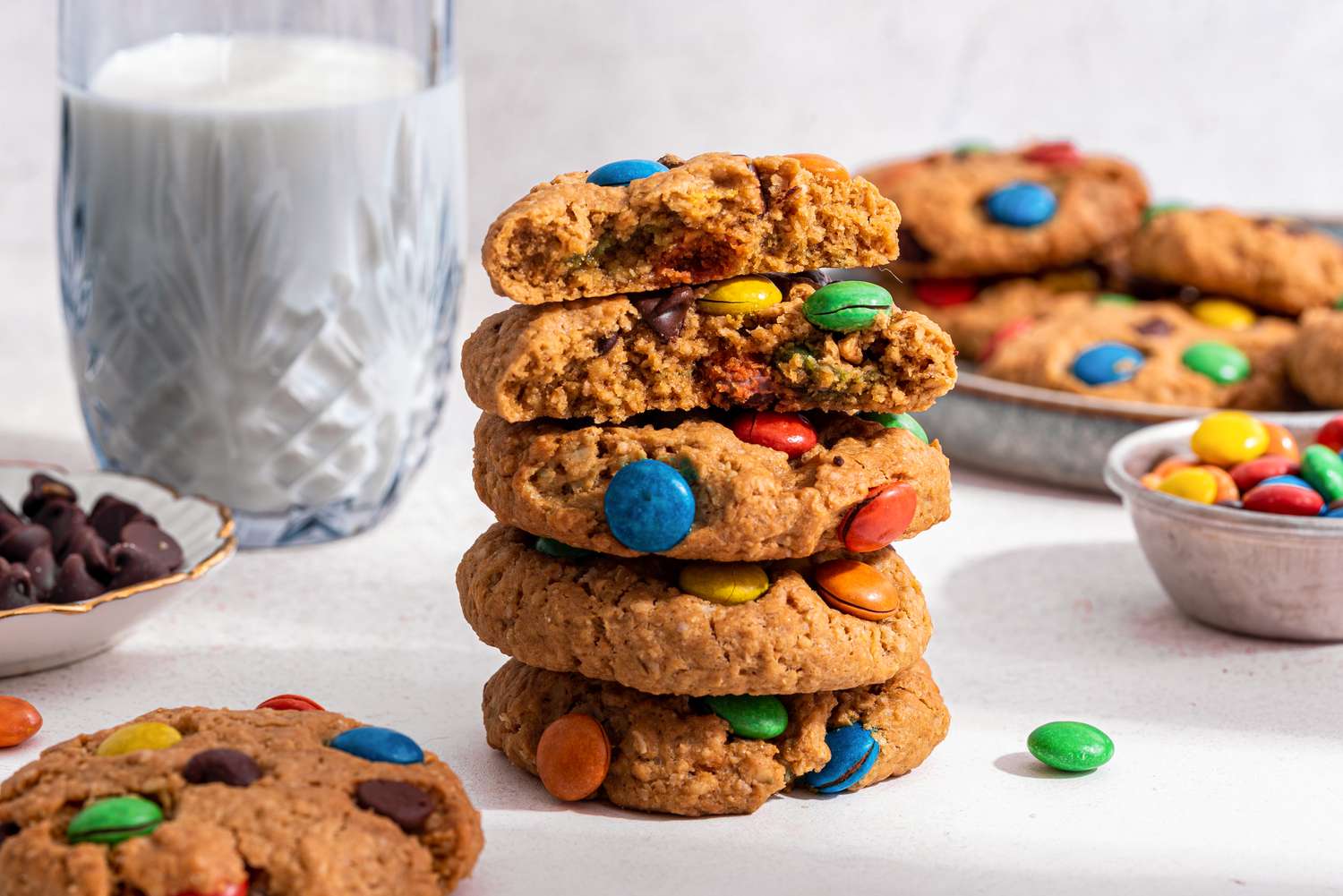 Stack of monster cookies with the top cookie broken in half, and in the background, a glass of milk, a plateful of monster cookies, a small bowl of chocolate chips, and a small bowl of M&M's 