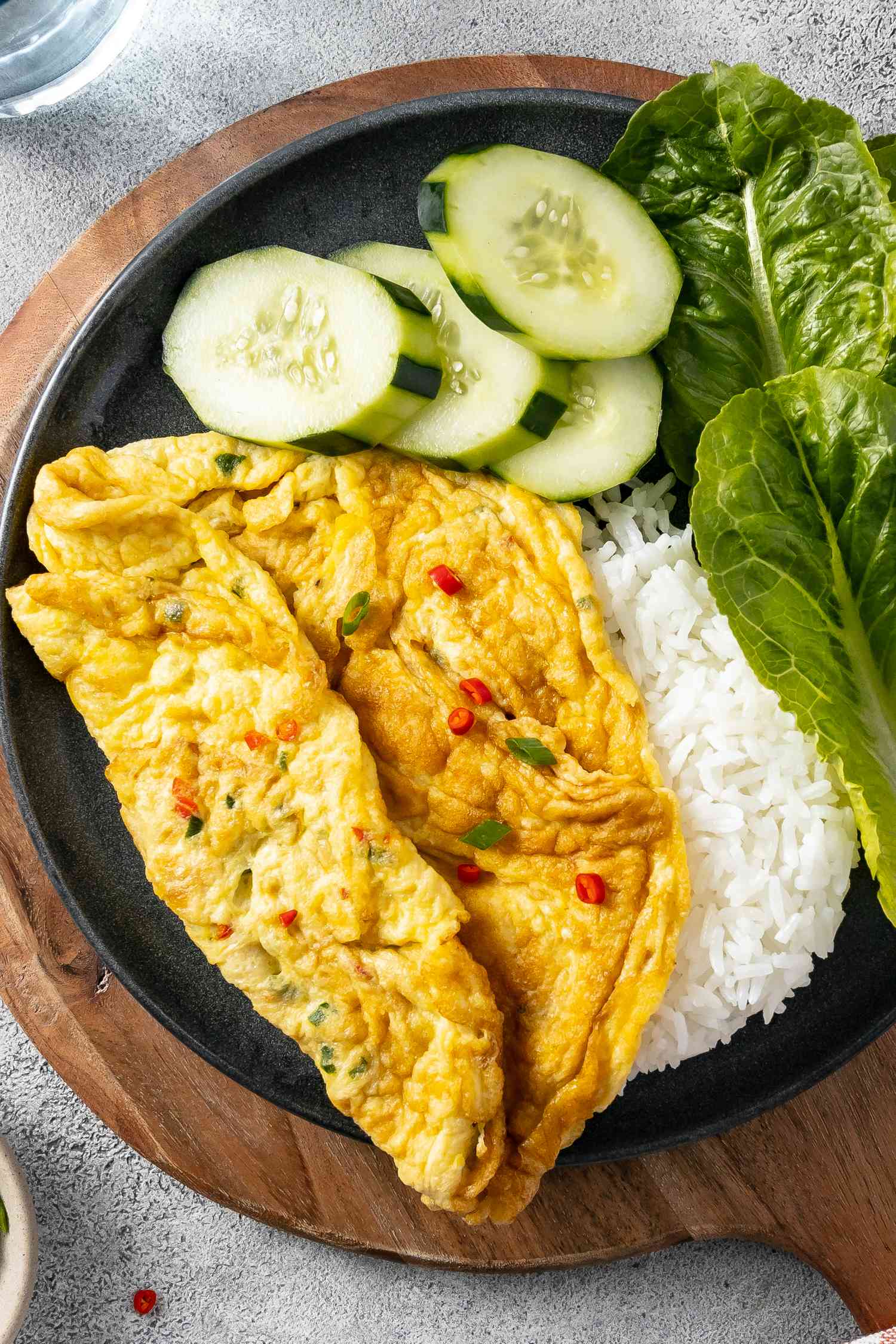 Plate of Kai Jeow (Thai Omelet) with Cucumber Slices, Romaine Leaves, Rice, and a Small Bowl of Sriracha on a Wooden Circular Serving Tray