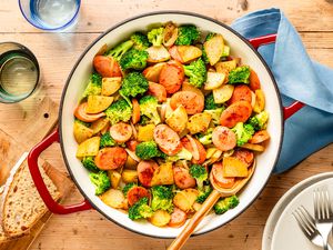 Overhead view of a red and white cast iron skillet of Kielbasa, Potato, and Broccoli Skillet with a serving spoon next to slices of bread on a cutting board, two drinking glasses and a blue fabric napkin