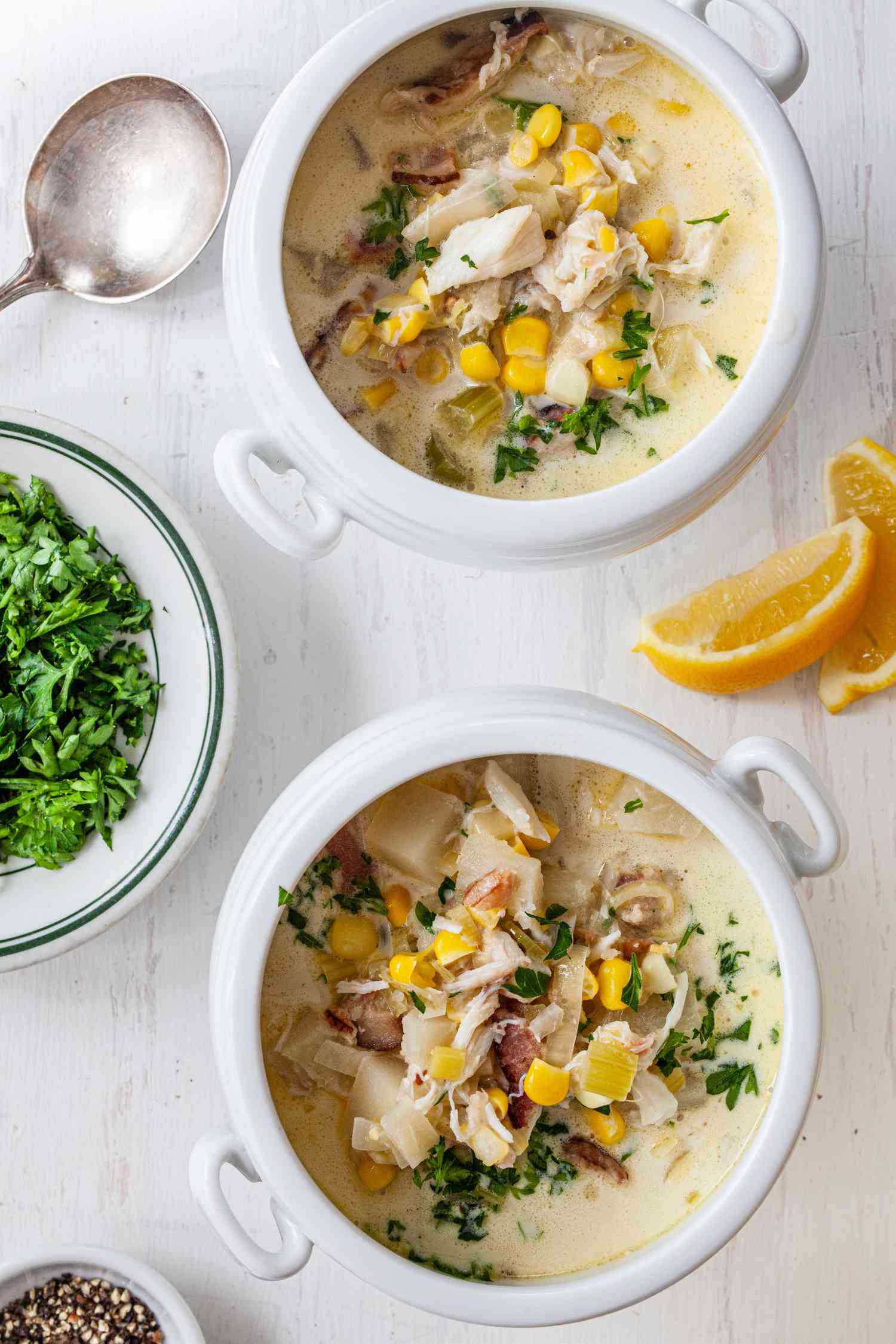 Corn and Crab Chowder in Two Soup Bowls with Handles at a Table Setting with a Plate of Parsley, Small Bowl of Cracked Pepper, and Lemon Slices on the Counter