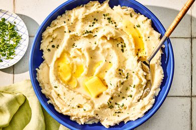 Overhead view of a large blue bowl of boursin mashed potatoes with a serving spoon and topped with herbs and butter all on a tile countertop