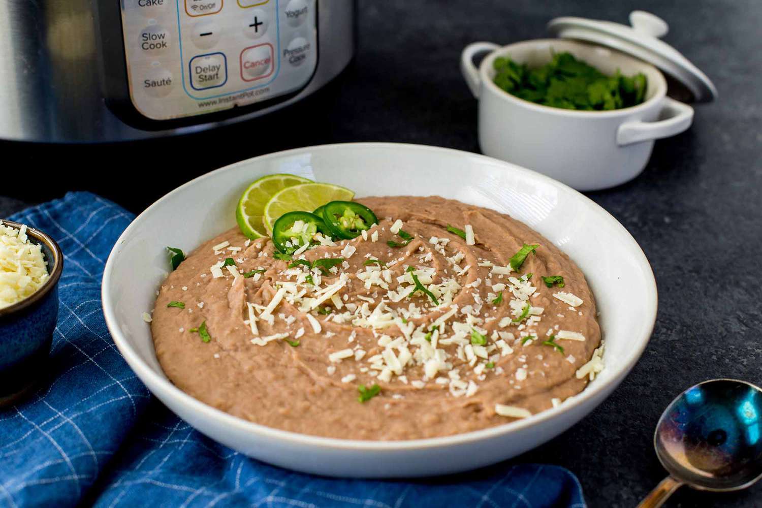 Homemade Refried Beans in a white bowl with lime wedges and sliced jalepenos nearby. The instant pot is in the background.
